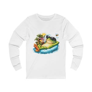 May include: White long-sleeve shirt featuring a cartoon frog wearing sunglasses and relaxing on a blue inflatable raft. The frog is holding a tropical drink, with palm trees, a sun, and a bee in the background.