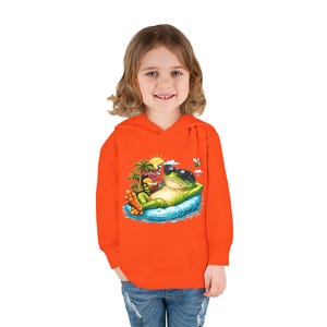 May include: Orange hooded sweatshirt featuring a graphic of a relaxed frog wearing sunglasses, lounging on a blue inflatable raft. The frog is holding a tropical drink, with palm trees, a sun, and a bee in the background. The child is wearing blue jeans.