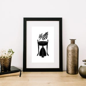 May include: Black and white framed art print featuring a stylized illustration of a decorative table with a plant. The table has a round top, curved legs, and a triangular base. A sprig of leaves sits in the table's bowl.
