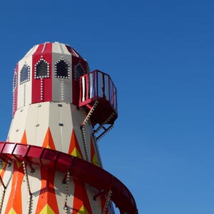 Può includere: Un'attrazione colorata della fiera con una parte superiore a strisce rosse e bianche, uno scivolo rosso e un balcone. La giostra presenta un design geometrico con accenti arancioni e gialli. Lo sfondo è un cielo blu chiaro.