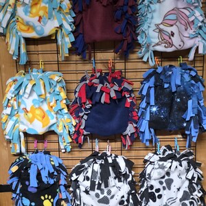 May include: A collection of fleece pet beds in various patterns and colors, including ducks, a unicorn, paw prints, and a galaxy design. Each bed has a fringed edge and is suspended from a wire grid.