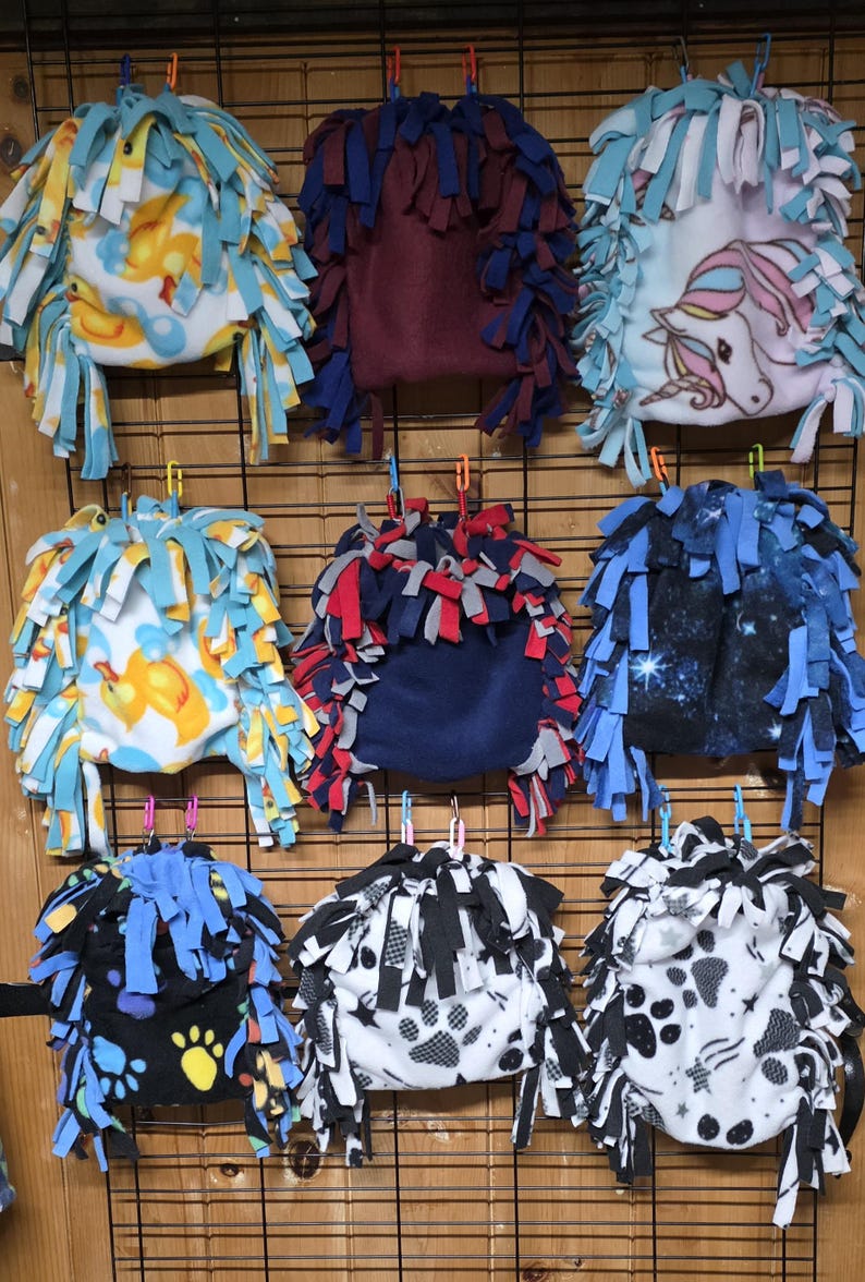 May include: A collection of fleece pet beds in various patterns and colors, including ducks, a unicorn, paw prints, and a galaxy design. Each bed has a fringed edge and is suspended from a wire grid.