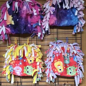 May include: Four fleece pet toys. Two have a galaxy print with pink, purple, and blue accents, and the other two have a red background with cartoon monster designs. Each toy has a fringe of colorful fabric strips.