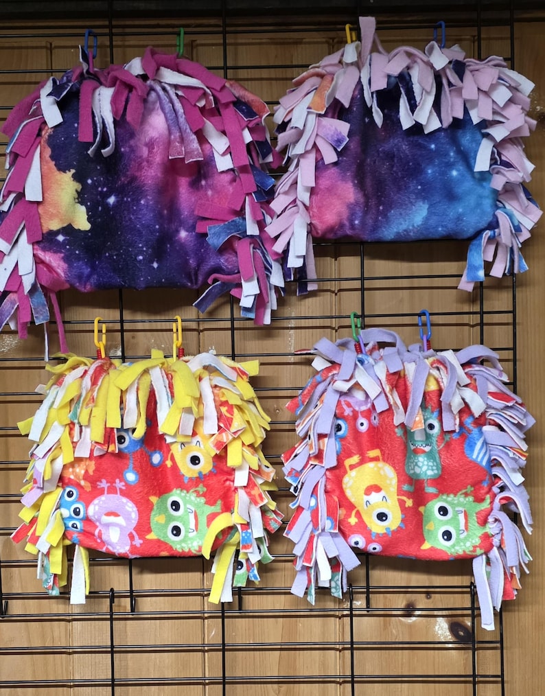 May include: Four fleece pet toys. Two have a galaxy print with pink, purple, and blue accents, and the other two have a red background with cartoon monster designs. Each toy has a fringe of colorful fabric strips.