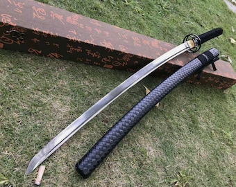 Puede incluir: Una espada katana japonesa con una hoja plateada, empuñadura negra y vaina negra. La espada descansa sobre hierba verde junto a una caja marrón con caracteres asiáticos.