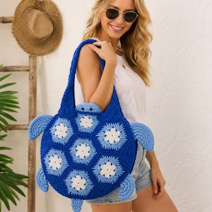 Könnte beinhalten: Eine gehäkelte Tragetasche in Form einer Meeresschildkröte mit einem tiefblauen Körper und hellblauen sowie weißen blumenartigen Sechseckmotiven. Die Tasche besteht aus einem Schildkrötenkopf, vier Flossen und einem Schwanz aus blauem Garn.