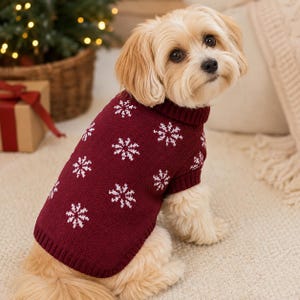 Puede incluir: Un perro pequeño de pelaje marrón claro lleva puesto un suéter de punto rojo oscuro con un patrón de copos de nieve blancos. El suéter tiene un cuello alto acanalado y puños acanalados en las aberturas de las patas para un ajuste ceñido.