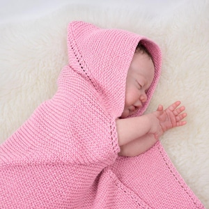 Op de afbeelding: Een zachte roze gebreide babydeken met een geïntegreerde capuchon en een verfijnde rand met gaatjespatroon. De deken is volledig in ribbelsteek gebreid, wat zorgt voor een eenvoudig en functioneel ontwerp voor baby's.
