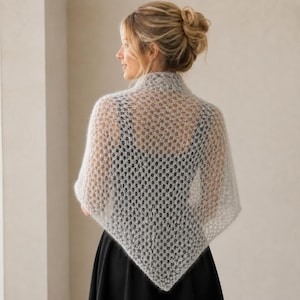 Puede incluir: Un chal triangular de punto en color gris claro, semitransparente, con un patrón abierto y repetitivo de panal. El chal está colocado sobre los hombros, mostrando una textura delicada y difusa con un dobladillo en punta hacia la espalda.
