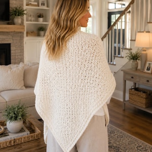 May include: A cream-colored triangular crochet shawl featuring a textured, open-stitch pattern. The shawl is draped over the shoulders, hanging down to a point at the back, and is paired with light-colored trousers, creating a neutral-toned ensemble.