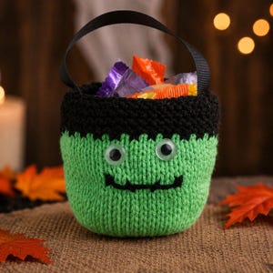 May include: A green knitted bucket with a black rim and a black ribbon handle. The bucket features two plastic googly eyes and a stitched black mouth, resembling a monster face. It is filled with various colorful wrapped candies.