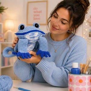 May include: A handmade crochet frog toy featuring a light blue body, a white patterned chest, large black eyes, and dark blue webbed feet. The frog has a wide mouth with a visible blue zipper closure running horizontally across the face.