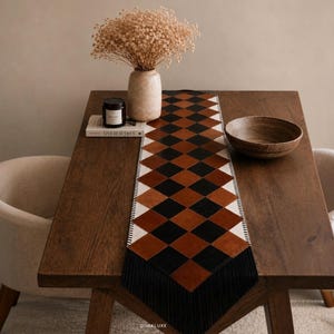 May include: A rectangular table runner featuring a repeating diamond pattern in alternating black, brown, and cream colors. The runner has a stitched border and black fringe at the ends, displayed centered on a dark wooden table surface.