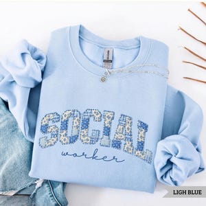 May include: A light blue crewneck sweatshirt featuring the word SOCIAL in a blue and yellow floral patchwork pattern, with the word worker written in cursive below. The garment is paired with blue denim jeans and a silver chain necklace.