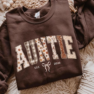 Puede incluir: Una sudadera de cuello redondo color marrón oscuro con la palabra AUNTIE en letras grandes, cada una rellena con un patrón otoñal diferente que incluye hojas, cuadros, calabazas y flores. Debajo del texto aparece en blanco EST. 2026 con un lazo.