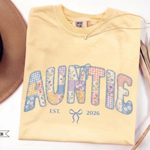 May include: A butter yellow crewneck t-shirt featuring the word AUNTIE in a patchwork-style font with floral and gingham patterns in blue, pink, and yellow. Below the text is the blue printed text EST. 2026 with a small blue bow graphic.