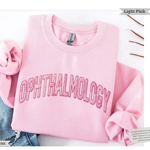 May include: A light pink crewneck sweatshirt featuring the word OPHTHALMOLOGY printed in a simulated pink and white gingham patchwork style across the chest. The garment has long sleeves and a ribbed collar, with a tag visible at the neckline.