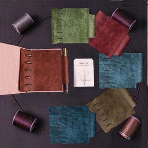 Può includere: Un set di copertine per quaderni in pelle scamosciata verde, rossa, verde acqua e marrone, con cinque fessure orizzontali e un passante per penna. Include un piccolo quaderno aperto con rilegatura ad anelli, una penna dorata, quattro rocchetti di filo e un biglietto con il testo THANK YOU for your kind support.