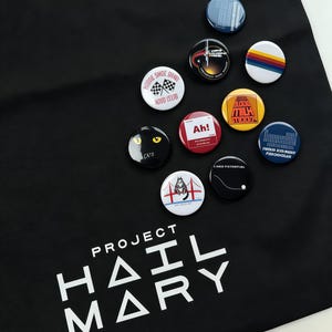 May include: A black fabric bag featuring the white text Project Hail Mary and a collection of circular pinback buttons. The buttons display various designs including a cat, a bridge, a checkered flag, and text such as Ah!, Blood Milk Moon, and Zero Potential.
