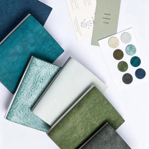 May include: A collection of rectangular notebooks with textured covers in shades of blue, green, and white, accompanied by a rectangular information card, a green card labeled NATURE RANGSAGE LIVING, and a white card with eight circular color swatches.
