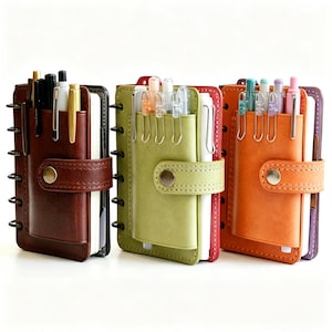 May include: Three small ring-bound planners in dark brown, olive green, and orange. Each planner features a front pocket with a snap button closure, containing multiple pens. The planners have visible metal rings along the side and stitched edges.
