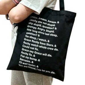 May include: Black tote bag with white text. The text includes phrases like "Amaze. Amaze. Amaze." and "You are friend, I no let you die." The bag is held by a person wearing a white t-shirt and gray sweatpants.