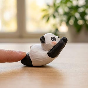 May include: A small, hand-carved panda figurine with black and white coloring. The panda is lying down with its arms raised, and has rosy cheeks. The figurine is on a wooden surface.