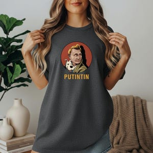 May include: Dark gray t-shirt featuring a graphic of a man with a dog inside a red circle. The text "PUTINTIN" is below the graphic in yellow letters. The shirt is a casual style.