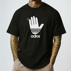 May include: Black t-shirt with a white graphic of a hand waving, stylized with three horizontal lines and the word "adios" below. The shirt is a short-sleeved crew neck.