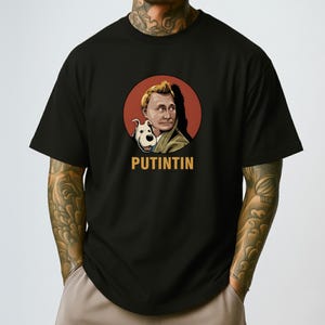 May include: Black t-shirt featuring a cartoon illustration of a man with a dog inside a red circle. The text "PUTINTIN" is printed in gold below the image. The shirt is made of a soft material.
