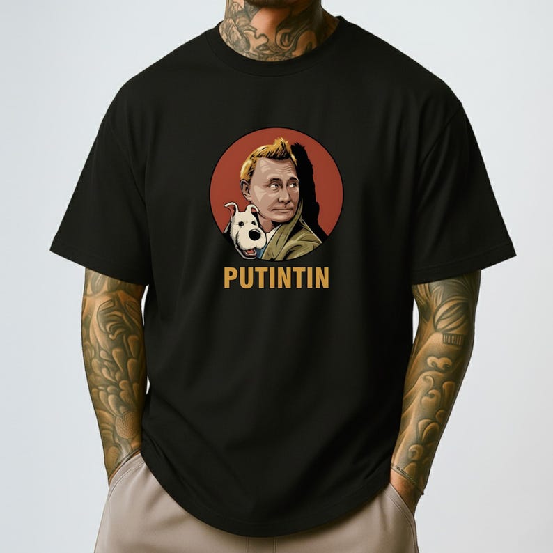 May include: Black t-shirt featuring a cartoon illustration of a man with a dog inside a red circle. The text "PUTINTIN" is printed in gold below the image. The shirt is made of a soft material.
