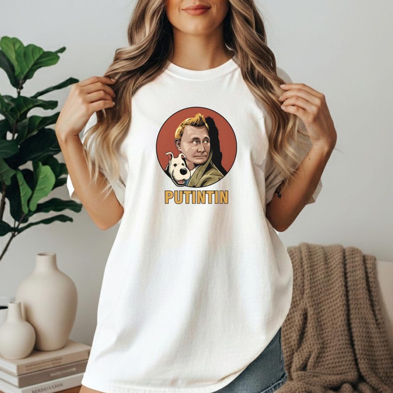 May include: White t-shirt featuring a graphic of a man and a dog inside a red circle. The text "PUTINTIN" is below the graphic in yellow letters. The shirt is a casual style.