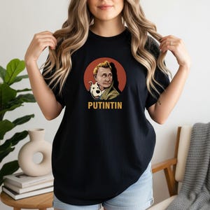 May include: Black t-shirt featuring a cartoon illustration of a man and a dog inside a red circle. The man has blond hair and is wearing a green jacket. The text "PUTINTIN" is printed in gold below the image.