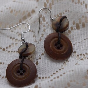 May include: A pair of button earrings. Each earring comprises a small, pale button above a larger, brown button, connected by dark string. The earrings hang from silver-coloured ear wires. The earrings are displayed on a white lace surface.