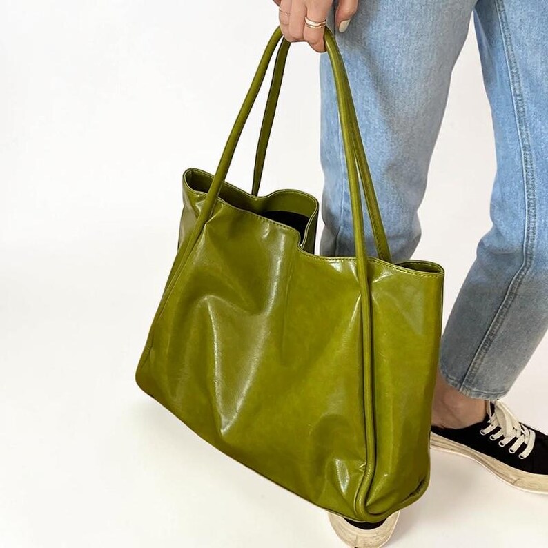May include: A large, olive green tote bag with long handles. The bag is made of a shiny, faux leather material. The bag is being held by a person wearing blue jeans and black sneakers.