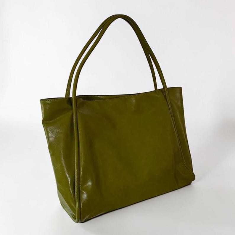 May include: Olive green tote bag with two long handles. The bag has a rectangular shape and a glossy finish. The handles are thin and made of the same material as the bag. The bag is empty and sits on a white background.