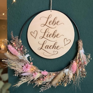 Könnte beinhalten: Ein runder Holzschild mit den Worten "Lebe Liebe Lache" und Herzsymbolen, aufgehängt an einem schwarzen Metallring. Der Ring ist mit getrockneten Blumen in Rosa, Beige und Weiß sowie kleinen Lichterketten verziert.