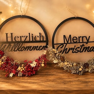 Könnte beinhalten: Zwei kreisförmige Wanddekorationen aus schwarzem Metall mit Kordelaufhängung. Das linke Schild trägt den Schriftzug Herzlich Willkommen mit roten und goldenen Trockenblumen. Das rechte Schild zeigt Merry Christmas mit silbernen und goldenen Trockenblumen.