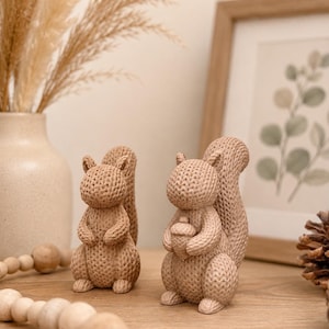 Op de afbeelding: Twee beige eekhoornbeeldjes met een gestructureerd, gebreid oppervlak. De ene eekhoorn staat rechtop, terwijl de andere een kleine eikel in de voorpootjes houdt. Beide figuren hebben gedetailleerde oren, ronde lichamen en pluizige staarten.