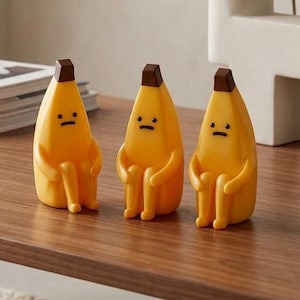 Könnte beinhalten: Drei gelbe, bananenförmige Figuren mit braunen Spitzen und kleinen schwarzen Gesichtern sitzen auf einem Holztisch. Die Figuren haben Arme und Beine und scheinen zu sitzen. Links befindet sich eine kleine Sukkulentenpflanze in einem grauen Topf.