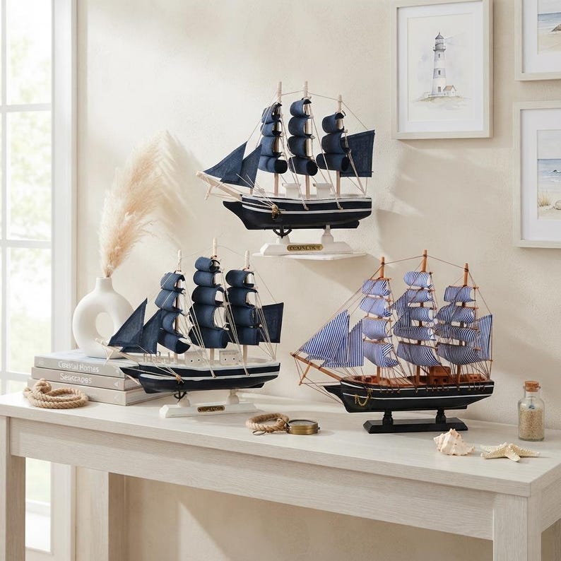 May include: Three model sailing ships displayed on a white table. Two ships feature dark navy blue fabric sails and black hulls, while the third ship has blue and white striped sails with a brown wooden hull. Each ship rests on a stand.