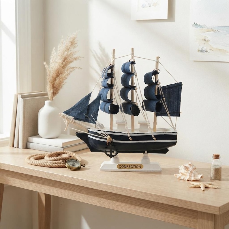 May include: A model ship with a black hull, white trim, and dark blue fabric sails. The ship features three masts, white rigging, and a small black anchor hanging from the bow. A white base displays the word CONFECTION in gold lettering.