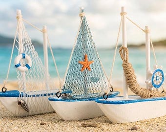 May include: Three decorative model sailboats on a sandy beach. The boats are white with blue trim and feature different nautical elements, including a life preserver, a starfish, and a ship's wheel. The sails are made of netting.