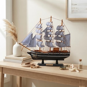 May include: A model sailing ship with a black hull, white horizontal stripes, and blue and white striped fabric sails. The ship features wooden masts, rigging, and a small plaque on the base labeled CONFECTION. It stands on a black pedestal.