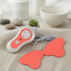 Könnte beinhalten: Ein weißes und korallenfarbenes elektronisches Gerät mit einer Reihe von Knöpfen und zwei korallenfarbenen Pads. Die Pads sind über ein weißes Kabel mit dem Gerät verbunden. Das Gerät verfügt über einen Ein-/Ausschalter, Lautstärkeregler und eine Wiedergabe/Pause-Taste.