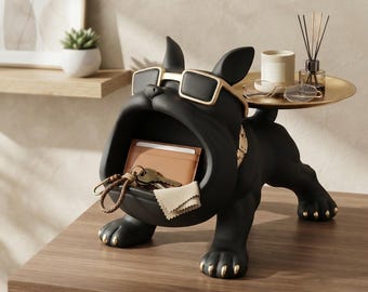 May include: A matte black bulldog figurine with gold-rimmed sunglasses and gold-tipped paws. The dog features an open mouth serving as a storage compartment for a tan wallet, keys, and a cloth, with a gold tray on its back holding a candle and glasses.