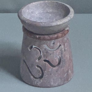 May include: A cylindrical stone oil burner with a mottled grey and brown finish. The top features a wide, shallow basin for oil, while the base has a cutout of the Om symbol, allowing light to pass through the carved stone structure.