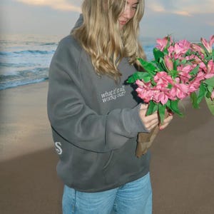 May include: A gray hoodie with the text "what if it all works out?" and a white "S" on the sleeve. The person is holding a bouquet of pink flowers. The background shows a beach and ocean.
