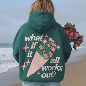 May include: A dark green hoodie with the phrase "what if it all works out?" in white text. The hoodie features a bouquet of pink flowers and stars. The person in the image is holding a bouquet of pink flowers.