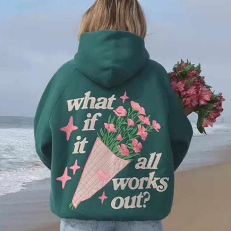 May include: A dark green hoodie with the phrase "what if it all works out?" in white text. The hoodie features a bouquet of pink flowers and stars. The person in the image is holding a bouquet of pink flowers.